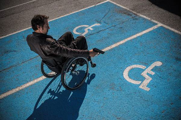 Rollstuhlfahrer steht auf Behindertenparkplatz und zielt mit Pistole auf Rollstuhlfahrersymbol