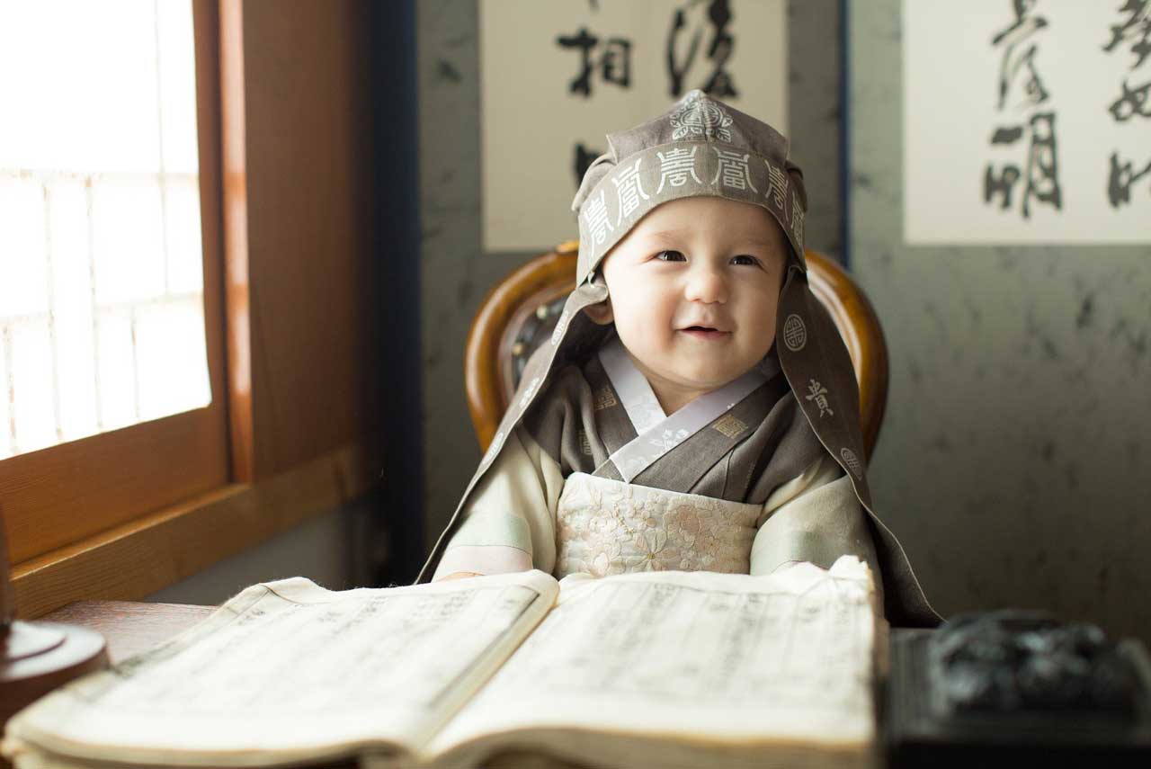 Baby in koreanischer Tracht sitzt vor aufgeschlagenem Buch.